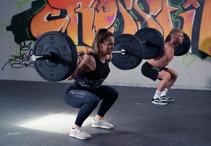 Kvinde og mand der udfrer squats med en vgtstang p ryggen.