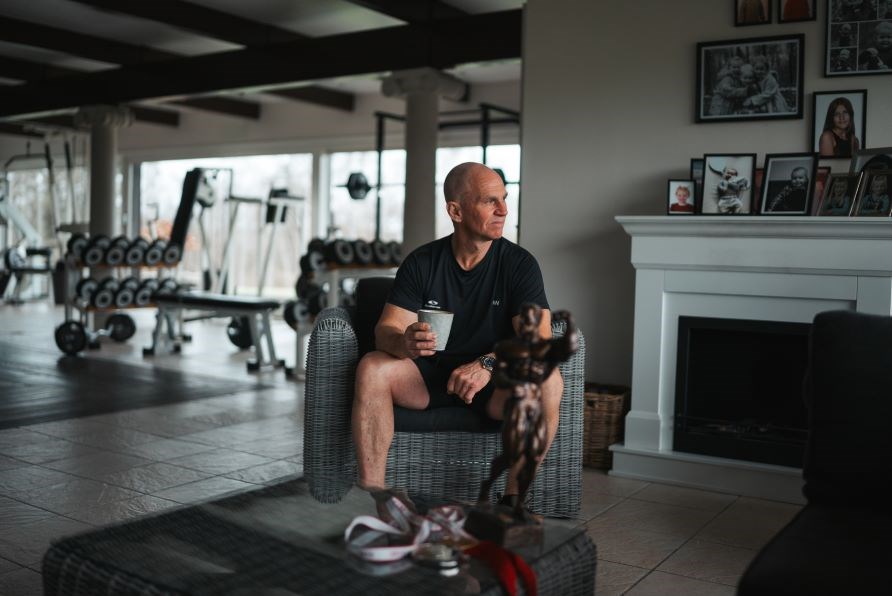 Michael Barnekow sidder i et hus i en stol med familiebilleder og trningsudstyr i baggrunden.