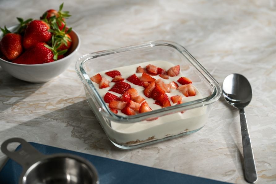 Glasmadkasse med proteinrig citron- og vaniljecheesecake, der st�r p� et bord sammen med en ske og en sk�l med jordb�r